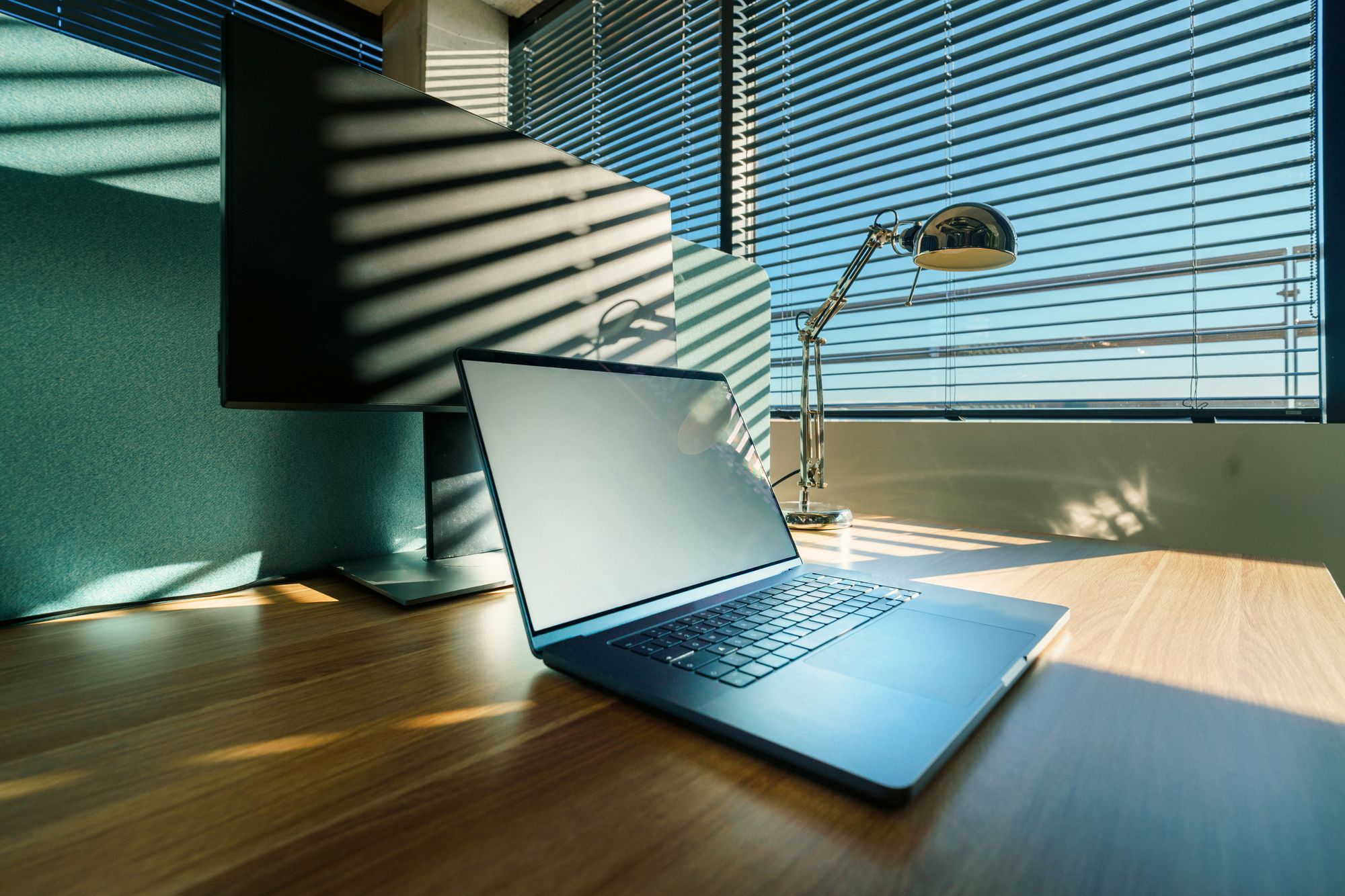 An empty desk with a laptop and a monitor beside window blinds. Reference photograph, not a LONG GAME VENTURES workplace.